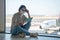 Female freelancer in medical mask sits in lotus position on airport floor waiting for flight to depart, and works using