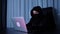 Female hacker sitting in dark room and typing on laptop keyboard. Woman in black mask and hood coding on screen and breaking passw