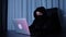 Female hacker sitting in dark room and typing on laptop keyboard. Woman in black mask and hood coding on screen and breaking passw