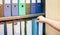 Female hand holding a binder of documents on the row of file folders that nicely management system on the office`s shelves