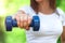 Female hand is holding a blue dumbbell against the background of the park. The girl is engaged in fitness in nature