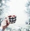 Female hand holding cup of tea with steam at snow nature landscape with snowfall. Polka dot cup with snowflakes. Mug of tea