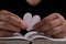 Female hand holding a half-torn white paper heart over an open book on the table. White paper heart torn in half in hand