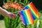 A female hand holding an LGBT rainbow flags against the backdrop of blooming Lilies
