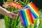 A female hand holding an LGBT rainbow flags against the backdrop of blooming Lilies