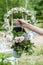 Female hand holds a bucket with lilac flowers, purple giant onions, and other flowers in front of a wedding arch