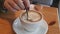 Female Hand Stirs Foam in Mug of Coffee by Spoon in Cafe Overlooking the Street