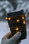 Female hand in winter warm gloves holding black eco paper cup with garland shiny light. Creative trendy zero waste