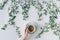 Female hands with a cup of coffee in branches with green leaves on a white table. Cofee art. Flat lay, top view.