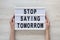 Female hands hold a lightbox with `Stop saying tomorrow` words over white wooden background, top view. Overhead, from above, fla