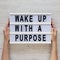 Female hands hold a lightbox with text `Wake up with a purpose` over white wooden background, top view. Overhead, from above, fl