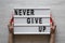 Female hands hold lightbox with words `Never give up` over white wooden surface, top view. Flat lay, from above, overhead