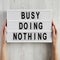 Female hands hold modern board with `Busy doing nothing` words on a white wooden background, top view. From above, flat lay,