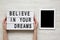 Female hands hold modern board with text `Believe in your dreams`, tablet wit blank screen over white wooden background. From ab