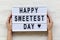 Female hands hold modern board with text `Happy Sweetest Day` word over white wooden background, top view. From above, flat lay