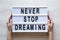 Female hands hold modern board with text `Never stop dreaming` over white wooden background, overhead. Top view, from above