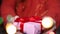 Female hands hold surprise gift box with red ribbon. Blurred garland in the foreground