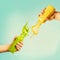 Female hands holding bottles with yellow and green splash smoothie or juice on blue background with tropical leaves and fruits