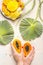 Female hands holding halved papaya fruit on white table with tropical leaves and plate with yellow sliced fruits, top view. Summer