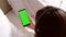 Female hands are holding a phone with a green screen. A girl with headphones listens to music