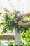 Female hands making vase with summer bouquet made from field wild flowers on a table in modern living room at window.