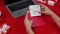 Female hands put greeting postcard with March 8th in festive envelope. Top view of a red table with a laptop and gift