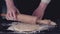 Female hands rolling out dough for pasta by wooden rolling pin over black kitchen table, powdering by flour. Dark rustic