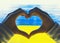 Female hands shaping heart symbol in background of yellow-blue flag of Ukraine on window. Human hands made heart shape on image of