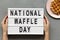 Female hans hold modern board with text `National Waffle Day`. Traditional belgian waffle on pink plate over concrete background