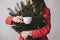 Female hiding his face behind a bouquet spruce branches, holding a cup of coffee on a grey background.