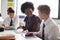 Female High School Teacher Sitting At Table With Students Wearing Uniform Using Digital Tablets In Lesson