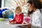 Female infant school teacher working one on one with a young white schoolboy, sitting at a table in a classroom writing, close up