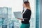 Female journalist writing an article using a laptop thinking standing at window and looking at city business district