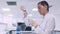 A female lab technician transfers a blue liquid sample to a test tube using a micropipette while sitting at a table in a