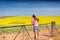 Female leaning on farm gate looks over rolling hills farmlands of golden canola