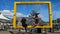 Female and male Tourists with face masks sitting on big yellow frame which frames table mountain. The man is a paraplegic and is