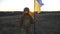 Female military medic walking at the field with waving flag of Ukraine. Ukrainian girl in uniform and helmet going with