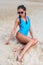 Female model with slender body wearing swimsuit sitting alone on sandy beach.