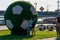 A female mother with a baby girl is hugging a huge green soccer ball. Tourists take pictures of her on a smartphone