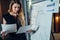 Female office worker working on her presentation standing near white board reading the report printed on paper