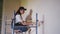 Female painter paints wall with roller on scaffolding at construction site. Young woman in overalls doing home