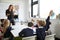 Female primary school teacher standing in a classroom gesturing to schoolchildren, sitting at a table raising hands