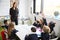 Female primary school teacher standing in classroom and schoolchildren sitting at table raising their hands