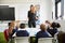 Female school teacher standing in a classroom gesturing to schoolchildren, sitting at a table listening