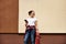 Female skateboarder with wooden red skateboard holding smartphone, enjoying listening to music posing outdoors near wall