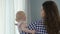 Female standing by window holding infant, child looking confused turning around