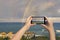 Female taking picture on mobile phone of double rainbow over ocean and tropical beach with umbrellas chairs and tables