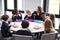 Female teacher kneeling to talk to a group of primary school kids sitting together at a round table eating their packed lunches