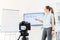 Female teacher is recording classes on a camera, she stands near flip chart and screen with a marker in hand, camera in foreground