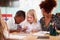 Female Teacher With Two Elementary School Pupils Wearing Uniform Using Digital Tablet At Desk